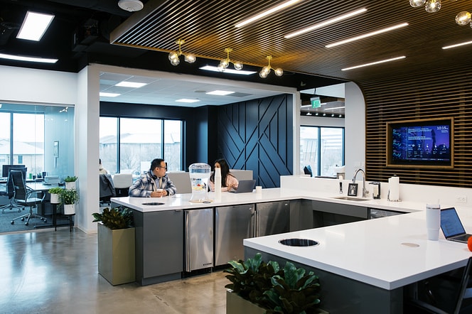 Open office café area with coworkers talking at a central island and large windows in the background