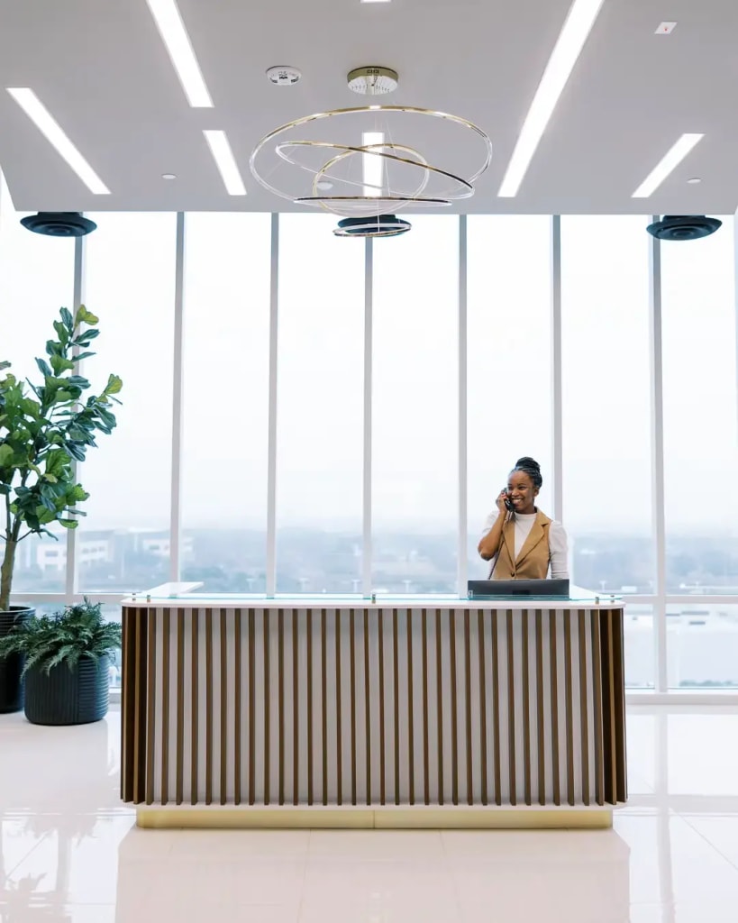 Reception: Lucid Private Offices reception desk with wood-slat front, gold ring chandelier, and panoramic city views from floor-to-ceiling windows.
