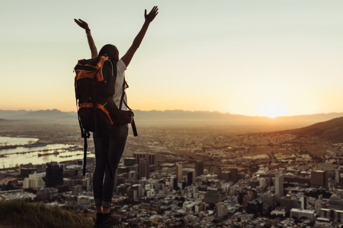 Backpacker girl on a lookout point over a city rising hands 