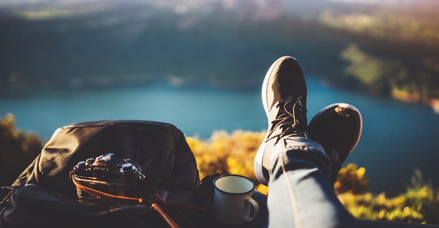 Imagem de pés, uma mochila e uma câmera e ao fundo a paisagem de um lago