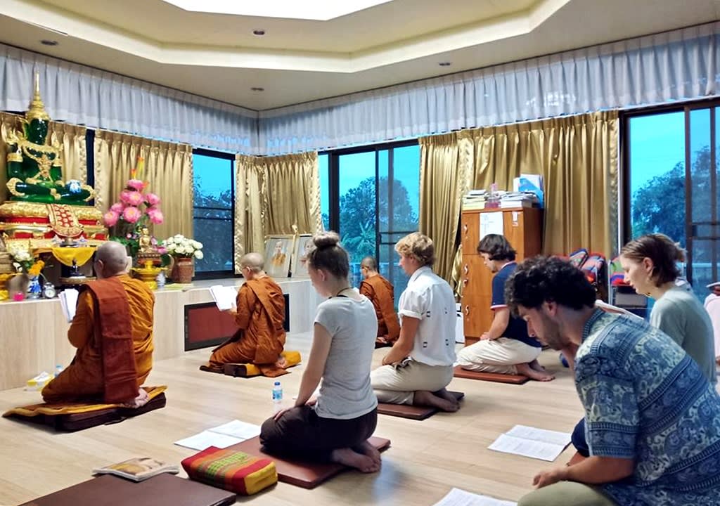 Western students reading mantras along with Buddhist monks facing a statue of Buddha