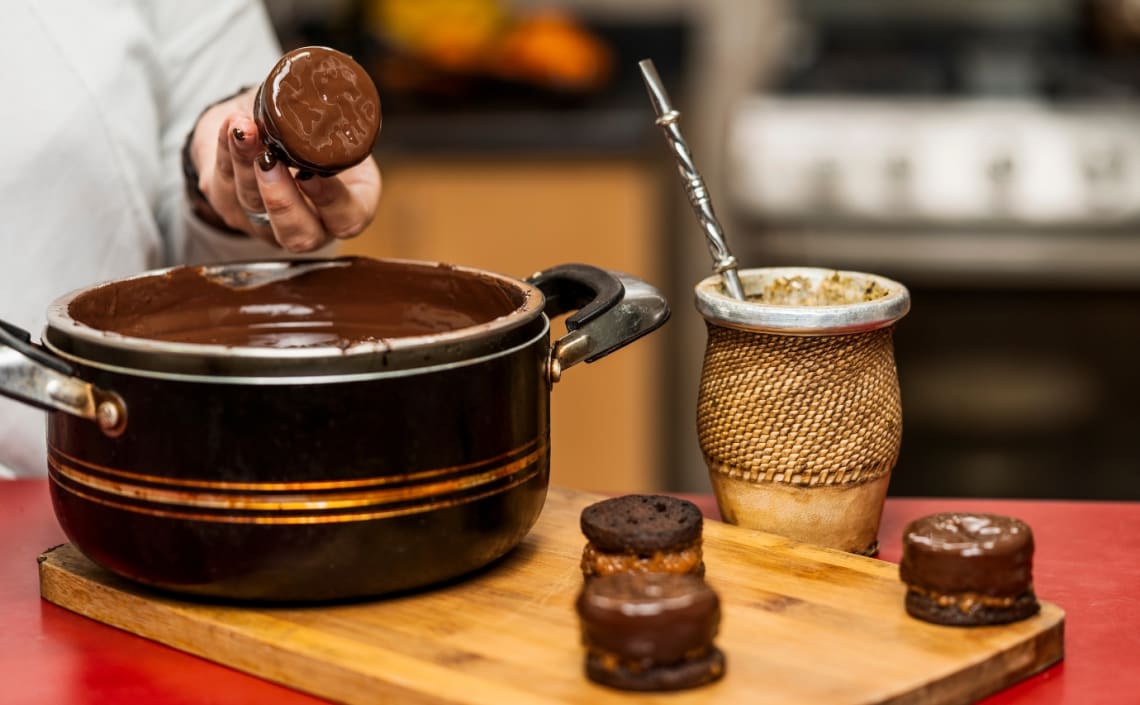 Mujer cubriendo alfajores en chocolate, y mate en la mesa