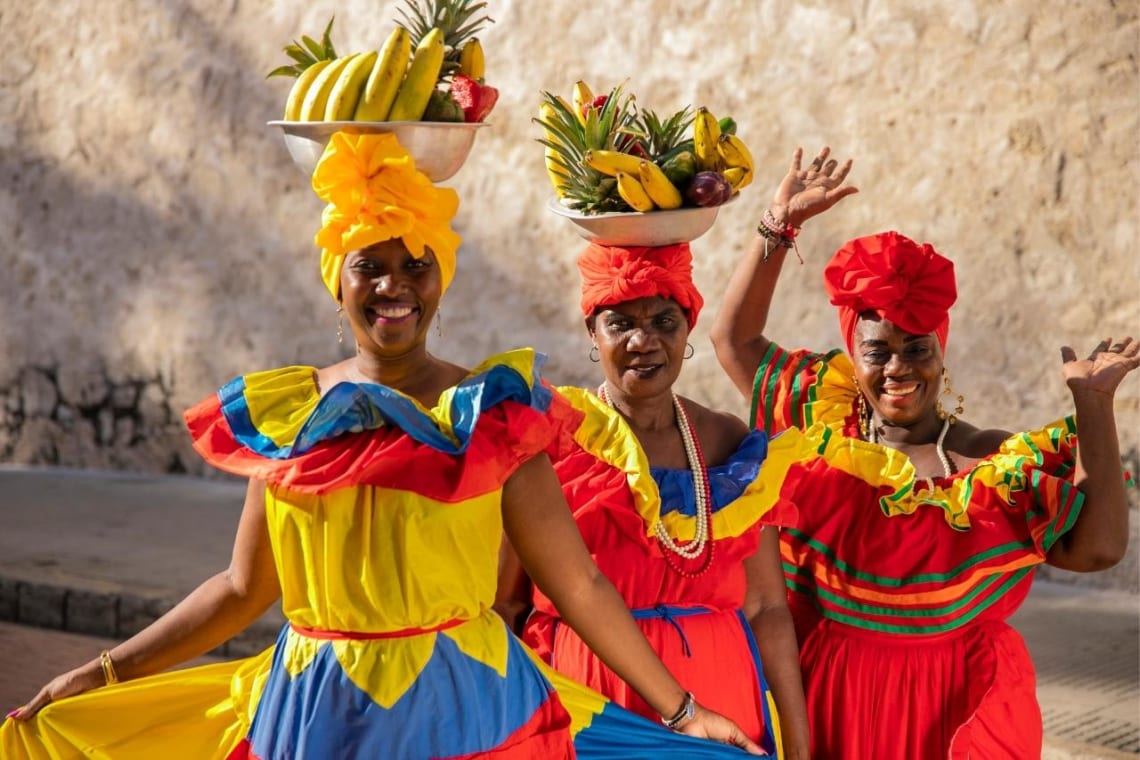 Three women in colorful traditional dresses from Colombia holding fruit bowls in their heads