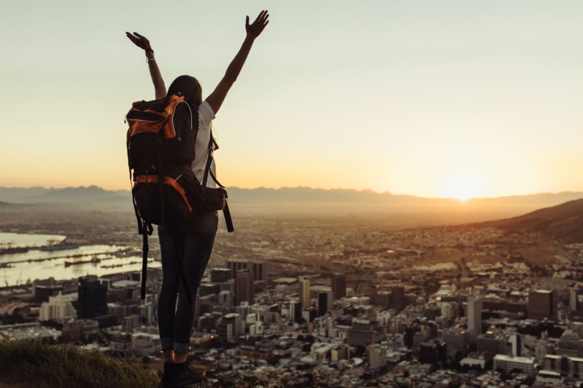Girl with a backpack rising his hand on a lookout overlooking a city at sunset