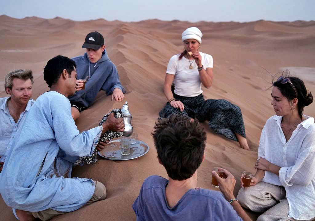 Three guys and two volunteer girls in a sand dune in Middle East dinrking tea with a local 