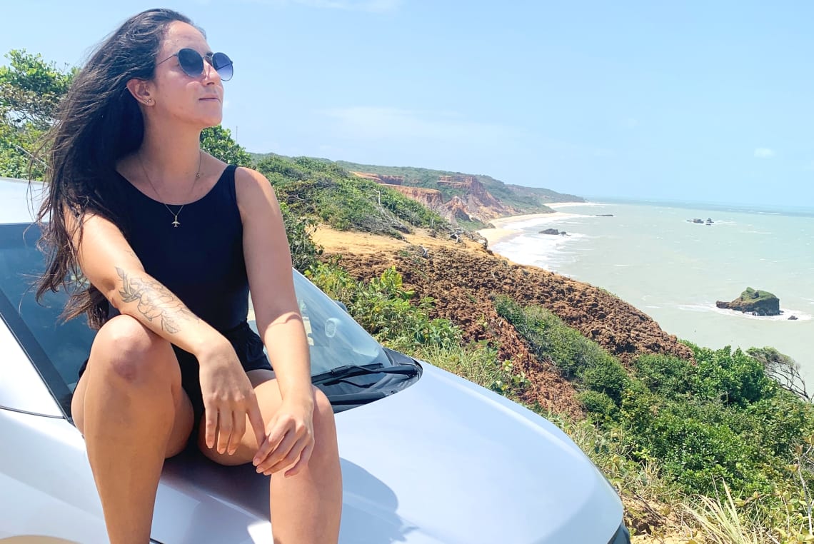 Mulher posando sentada no capô de um carro junto à praia em Conde