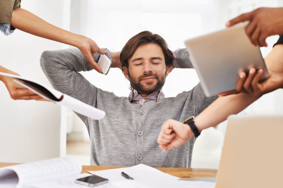 Man with eyes closed sorrounded by hands of workmates giving him a laptop, a phone and a planner