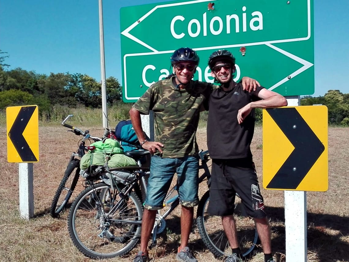 Padre e hijo ciclistas frente a cartel de la ruta que indica dirección para Colonia y Carmelo