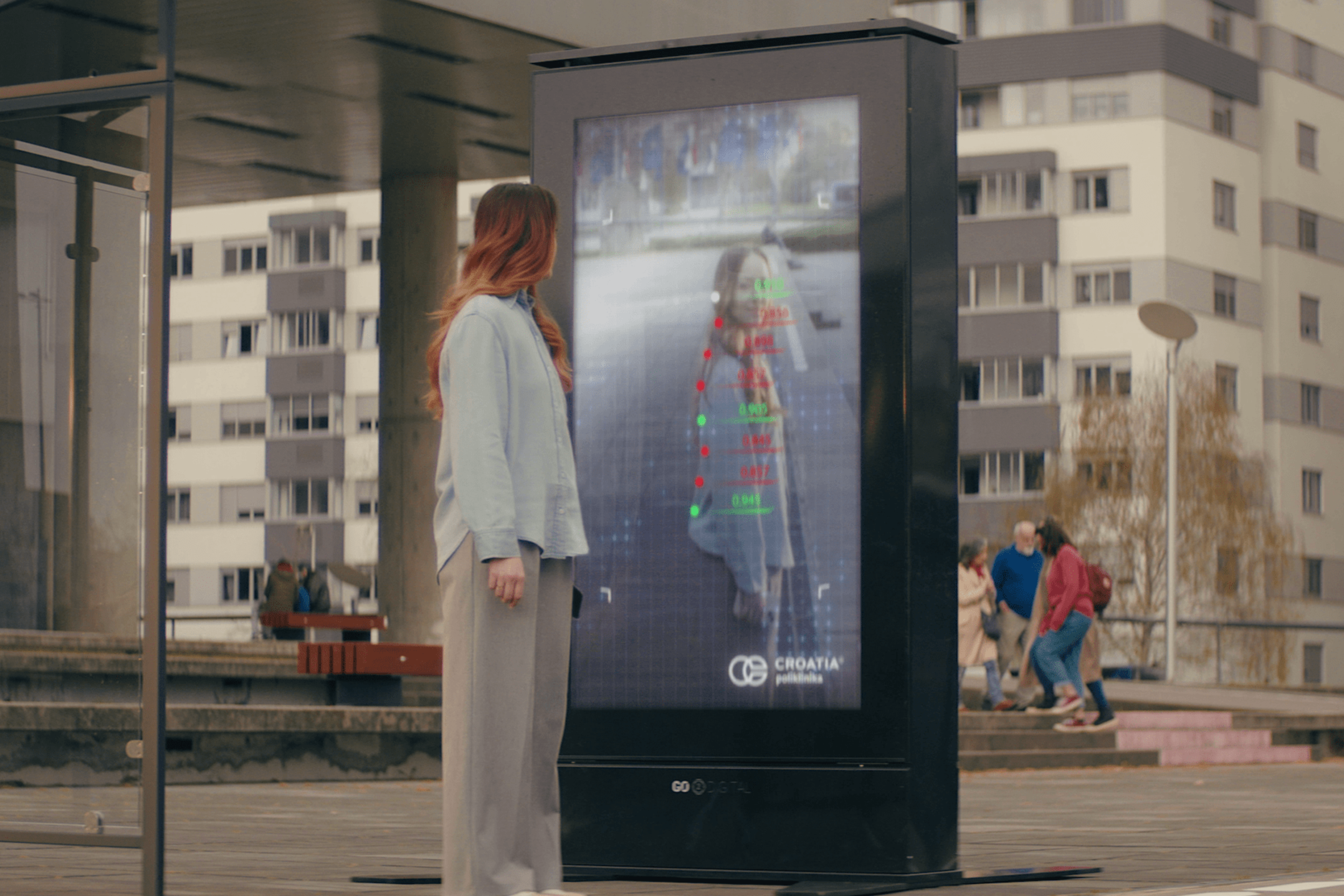 A woman stands in front of a digital advertising kiosk at a bus stop. The kiosk displays a live image of the woman with overlaid data points and the logo of "Croatia Poliklinika." In the background are apartment buildings and people walking.