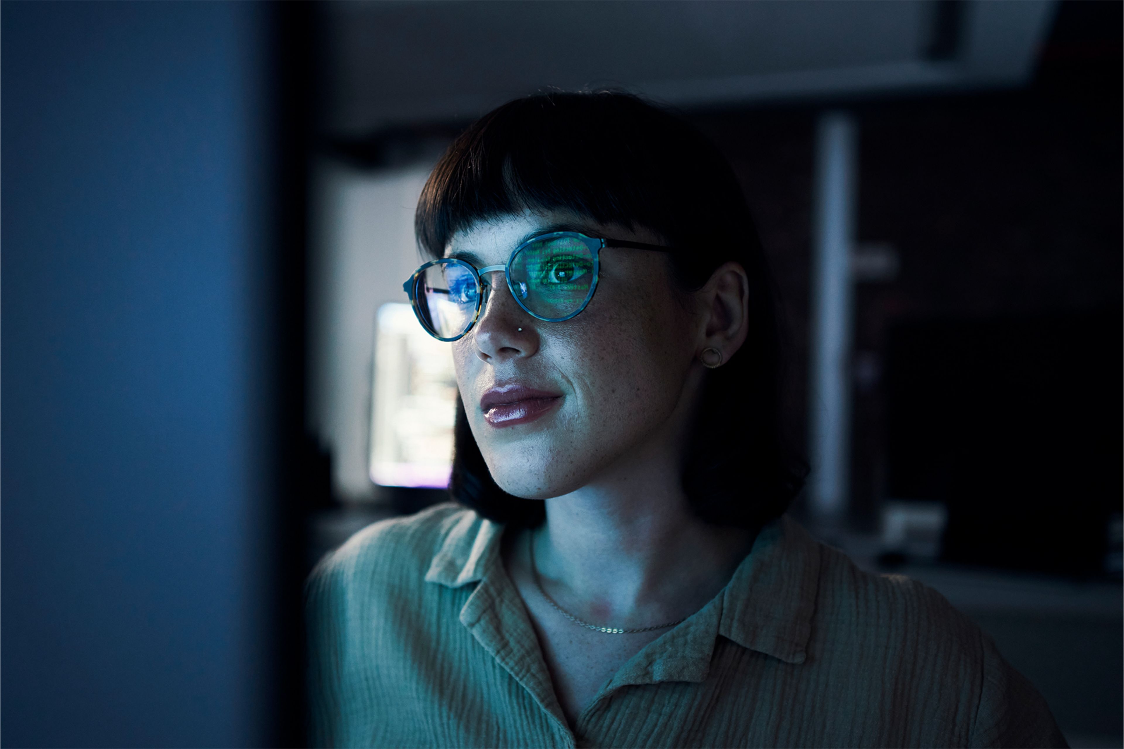 Person with short dark hair and glasses looking at a computer screen in a dimly lit room, with blue light reflecting on their face.