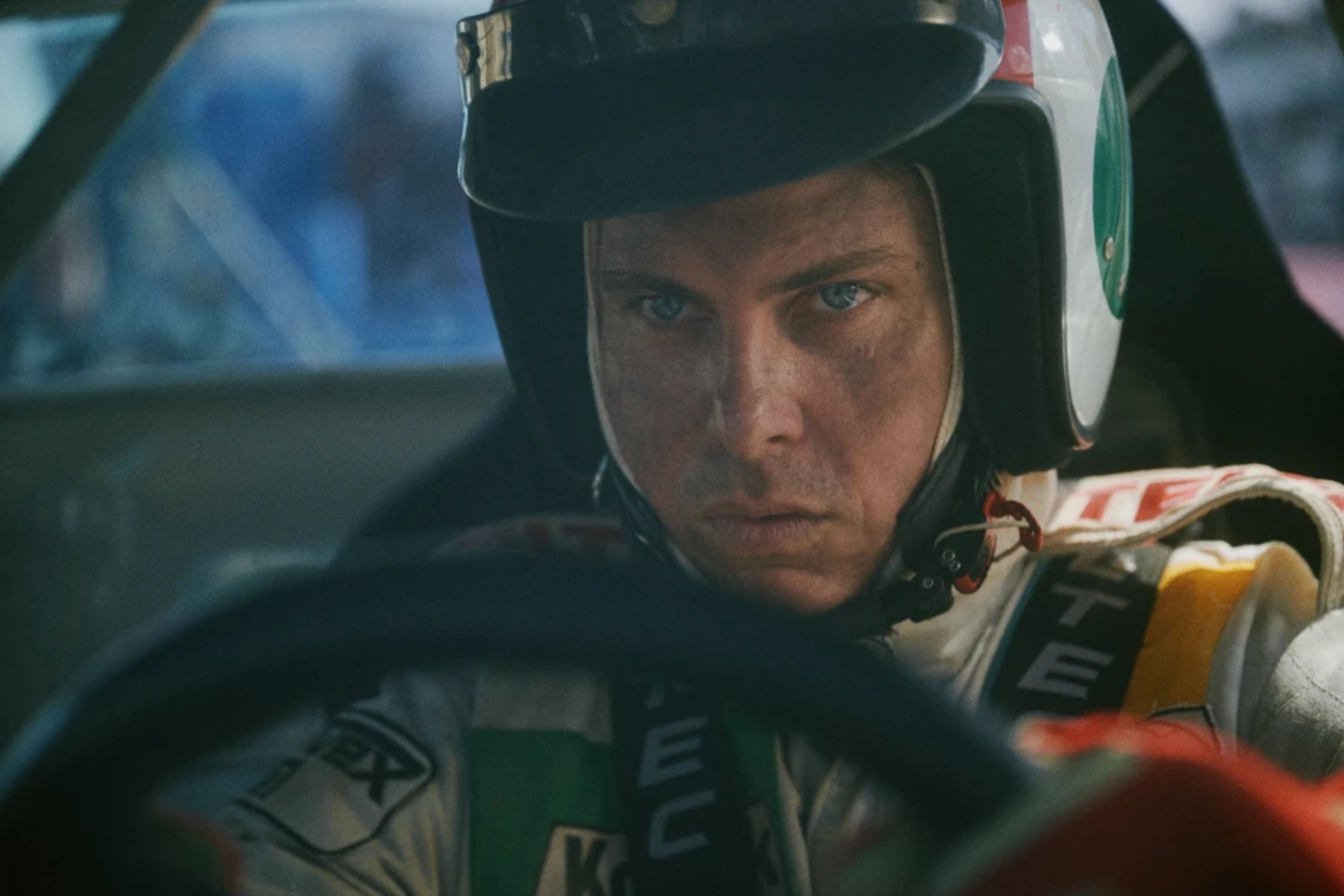 Close-up shot of a focused male racing driver behind the wheel, wearing a white and green helmet with a protective balaclava, staring intensely ahead from inside his race car.