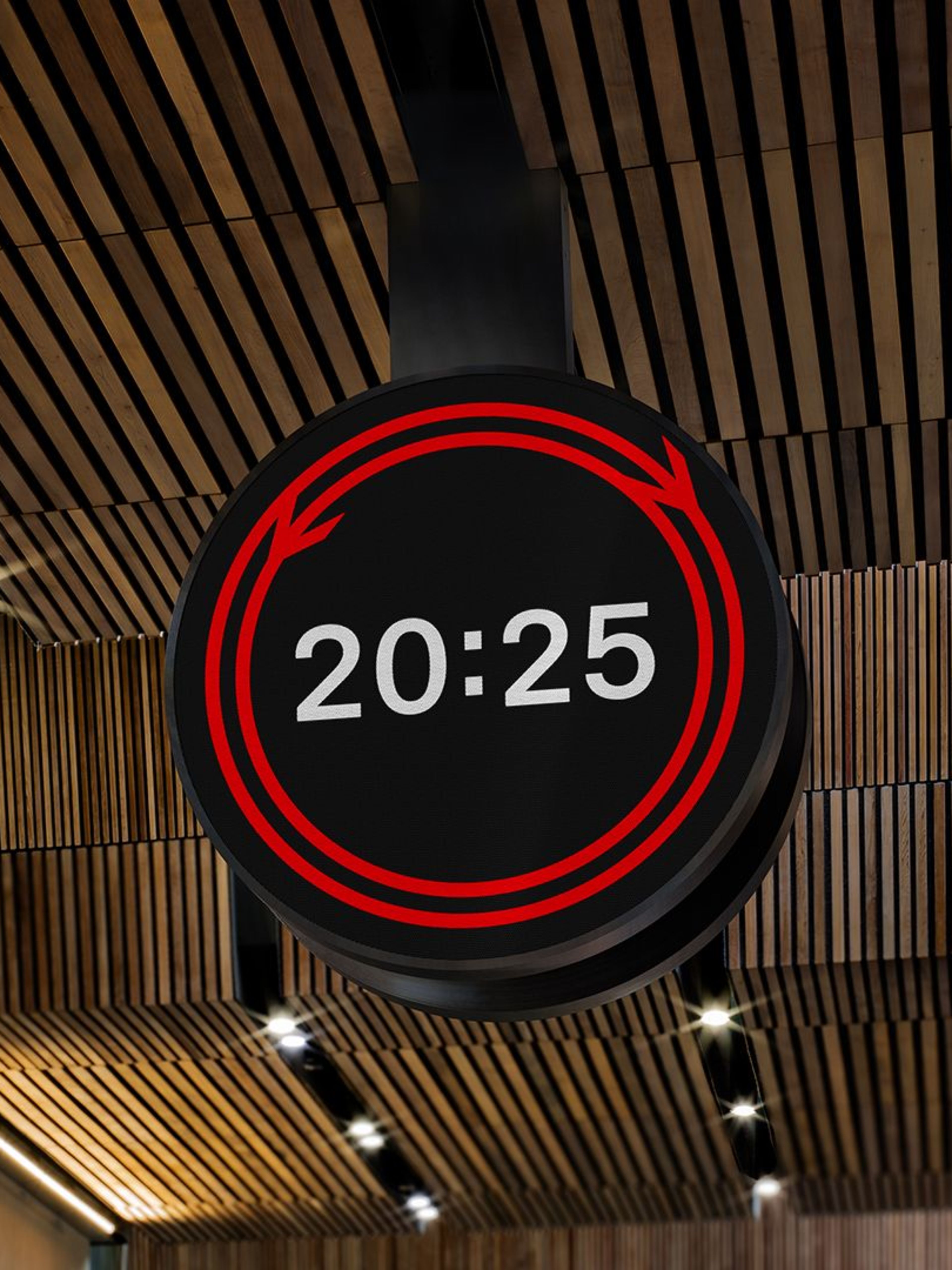 The circular Rail Clock hanging from a contemporary wooden-slatted station ceiling, featuring a digital display with a glowing red double-arrow graphic and white 24-hour time showing 20:25.