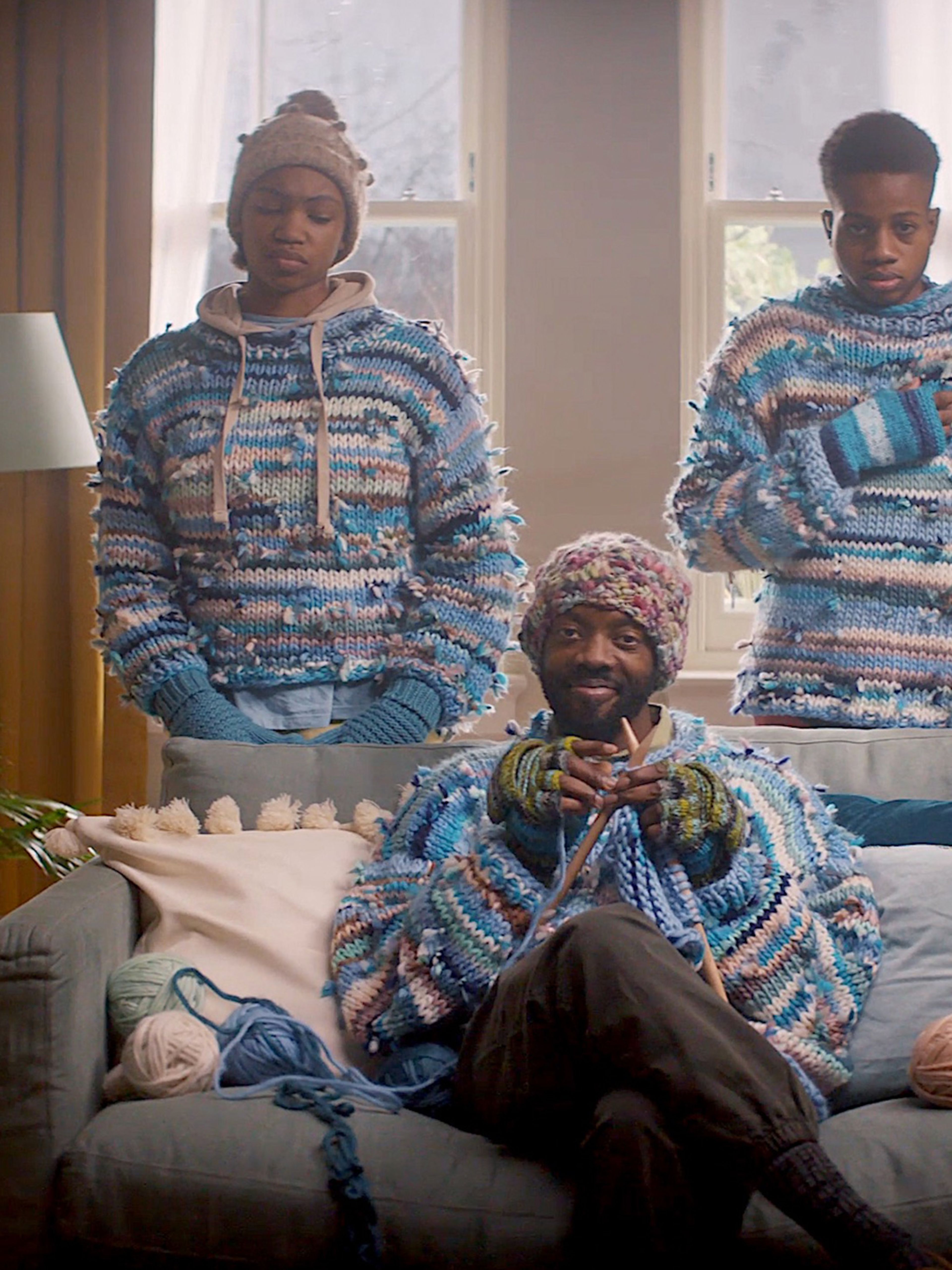 A father sitting on a sofa knitting with two teenagers standing behind him in hand knitted jumpers