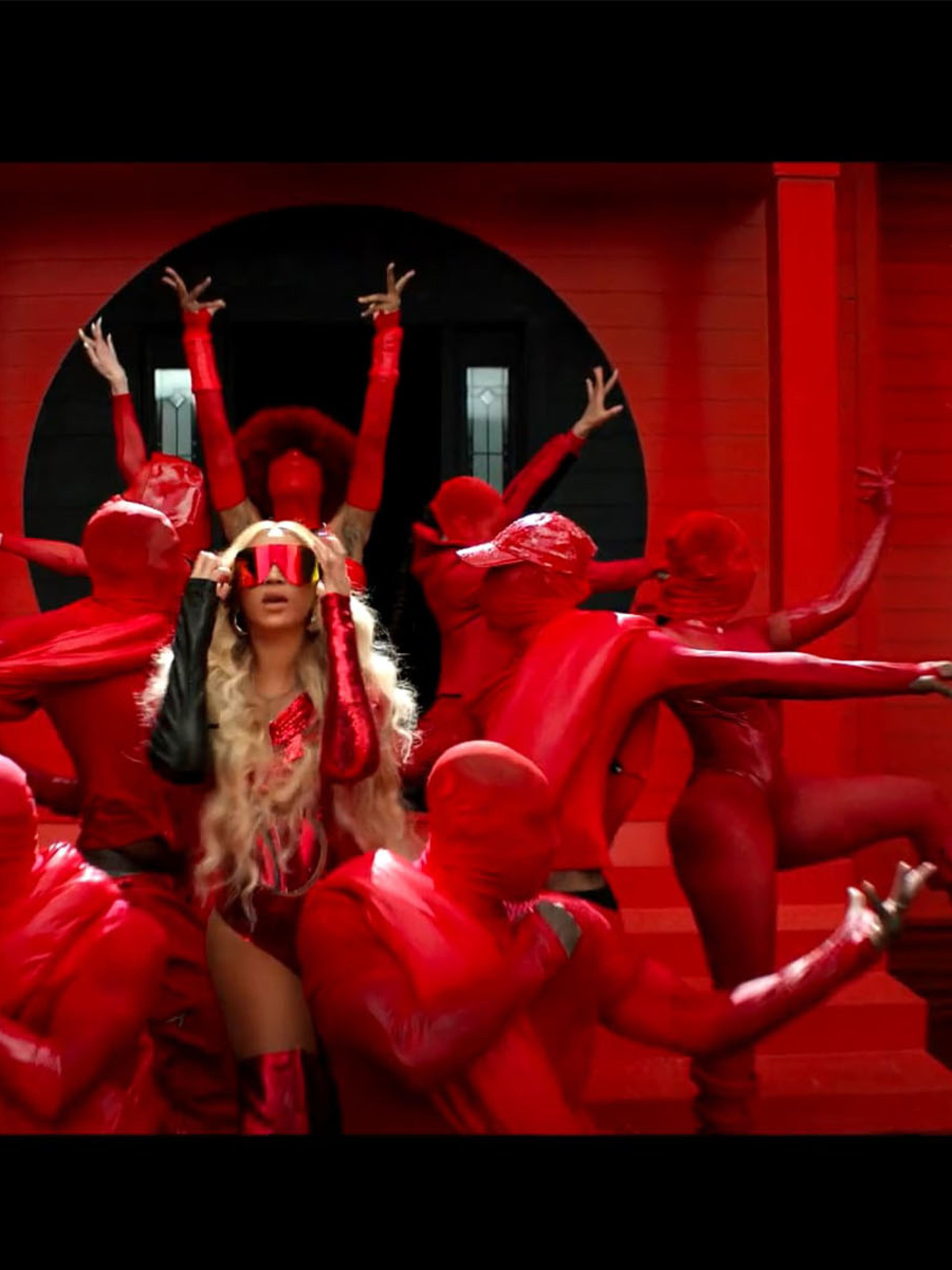 Beyonce dressed in red putting red sunglasses on her face, standing in front of a red house amidst a group on dancers also dressed in red
