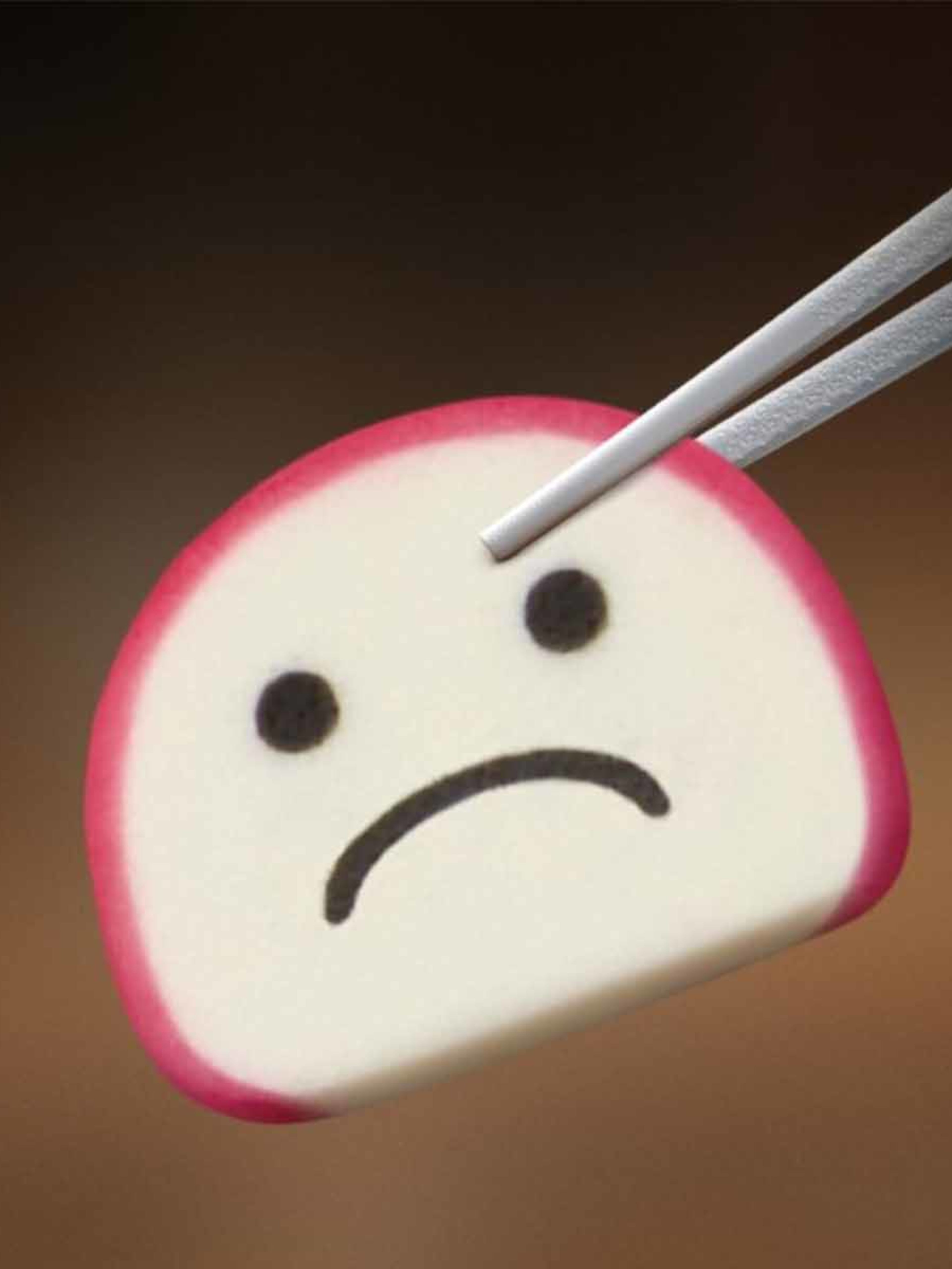 A close-up shot shows a piece of kamaboko (Japanese fish cake) being held by a pair of silver chopsticks against a blurred, dark brown background. The kamaboko is a semi-circular slice, white in the center with a thin, bright pink edge. On its white surface, a sad face is drawn in black: two small, round eyes and a downturned, curved mouth. The chopsticks are positioned at the top right, holding the kamaboko delicately.