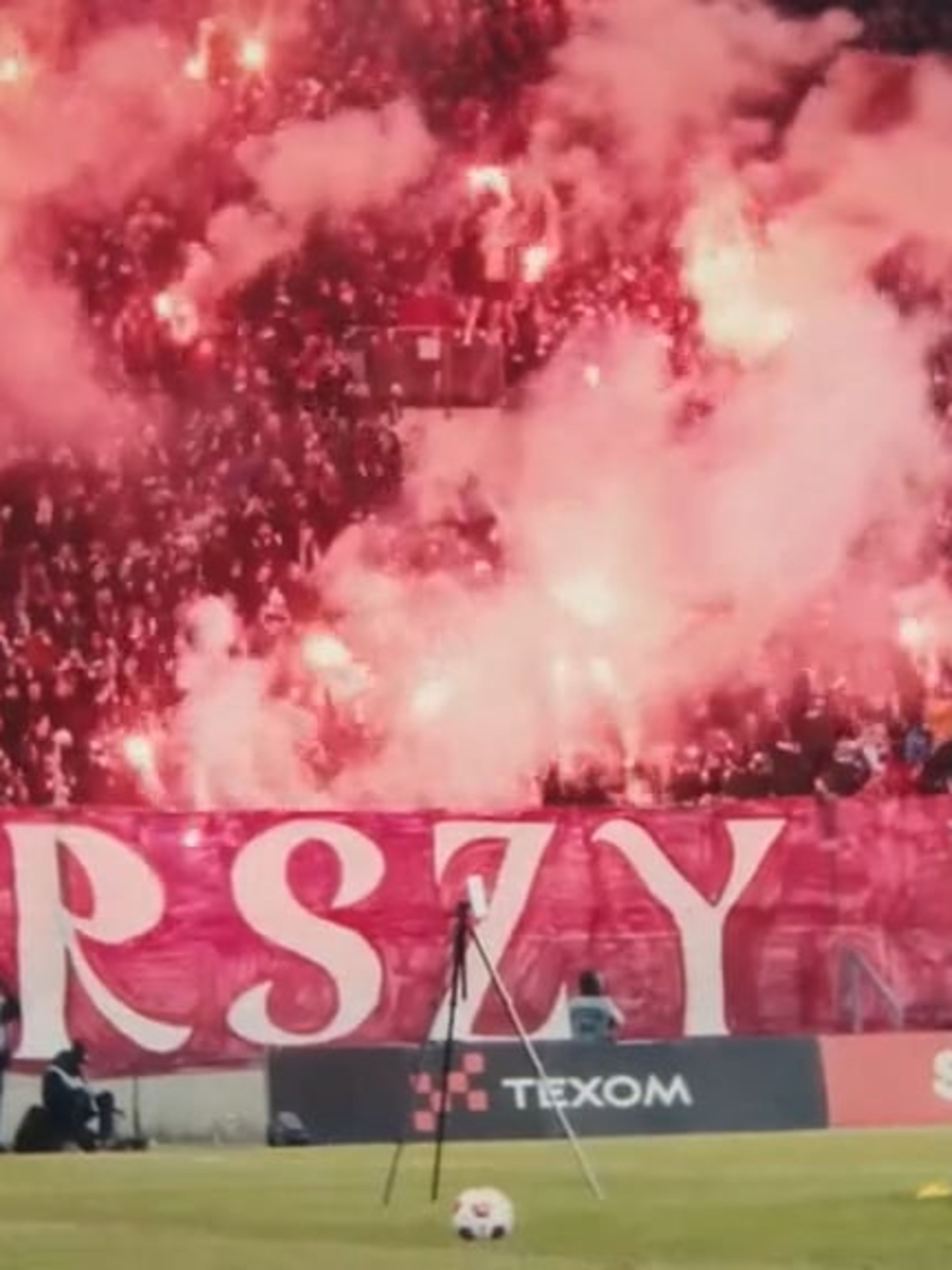 A packed stadium stand filled with Wisła Kraków football fans celebrating with red flares, creating a thick cloud of pink and white smoke. In front of the stand, a large red banner reads “STARSZY POLS” with advertising boards for Texom and Superbet visible at the edge of the pitch.