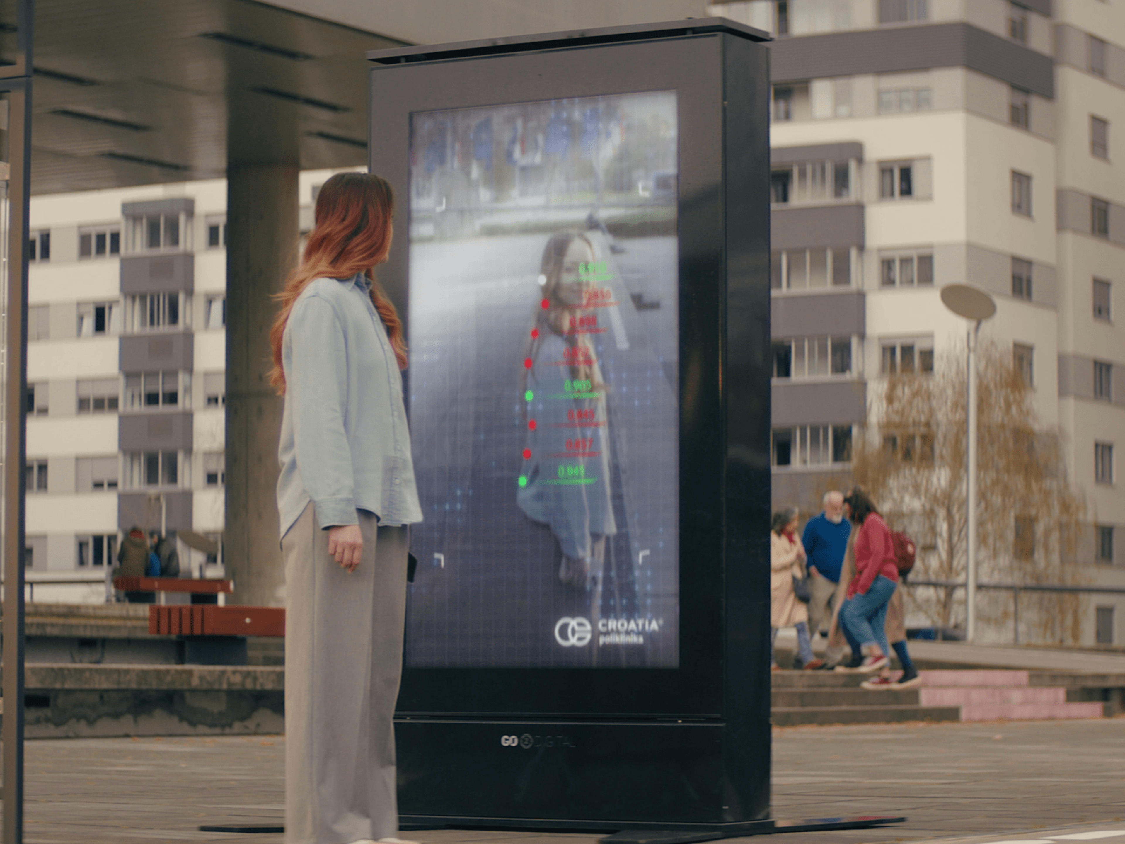 A woman stands in front of a digital advertising kiosk at a bus stop. The kiosk displays a live image of the woman with overlaid data points and the logo of "Croatia Poliklinika." In the background are apartment buildings and people walking.