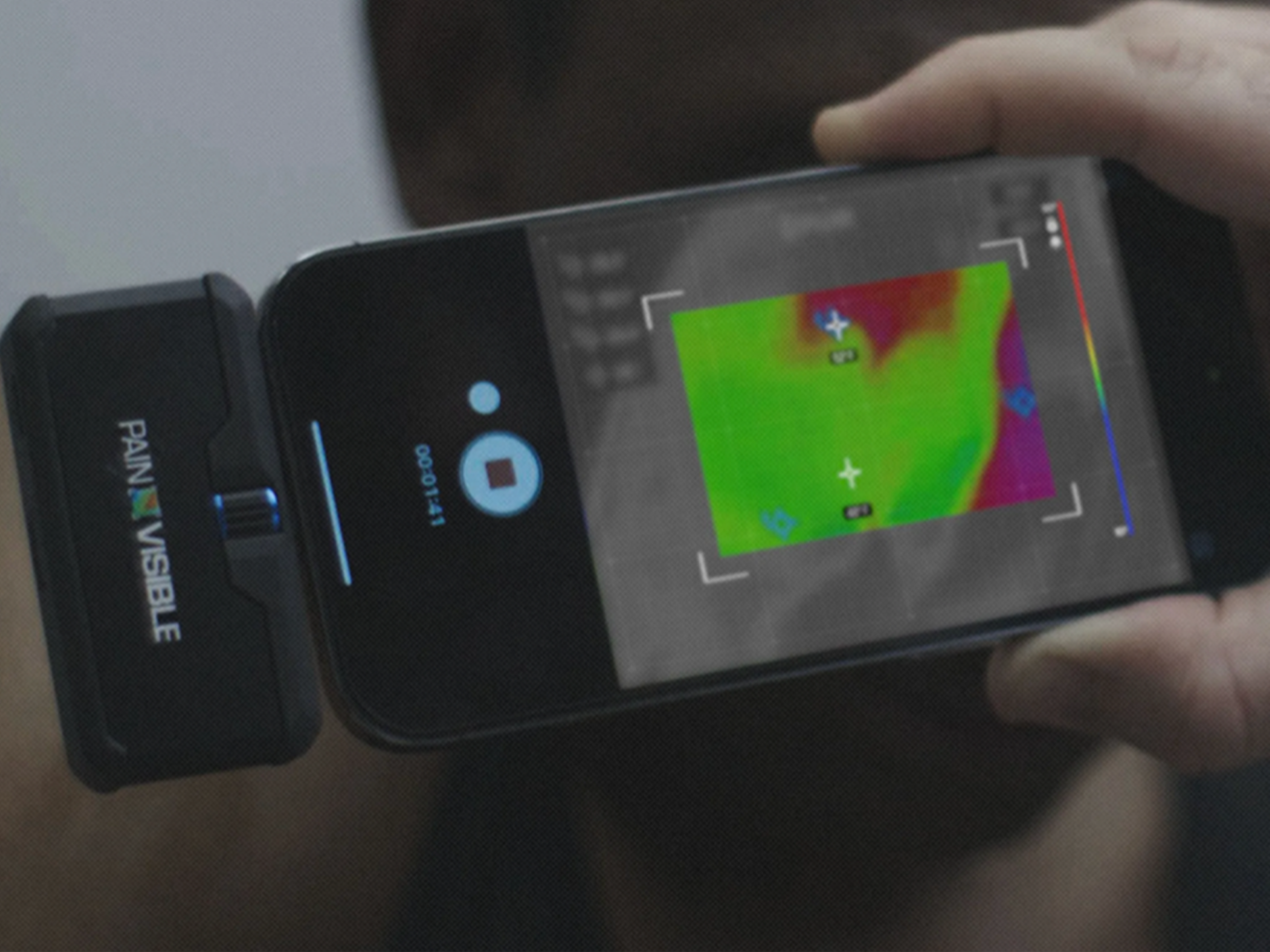 AI A person holds a smartphone horizontally with a "PAIN VISIBLE" attachment. The phone screen displays a thermal image in green and red, with crosshairs and a color scale.