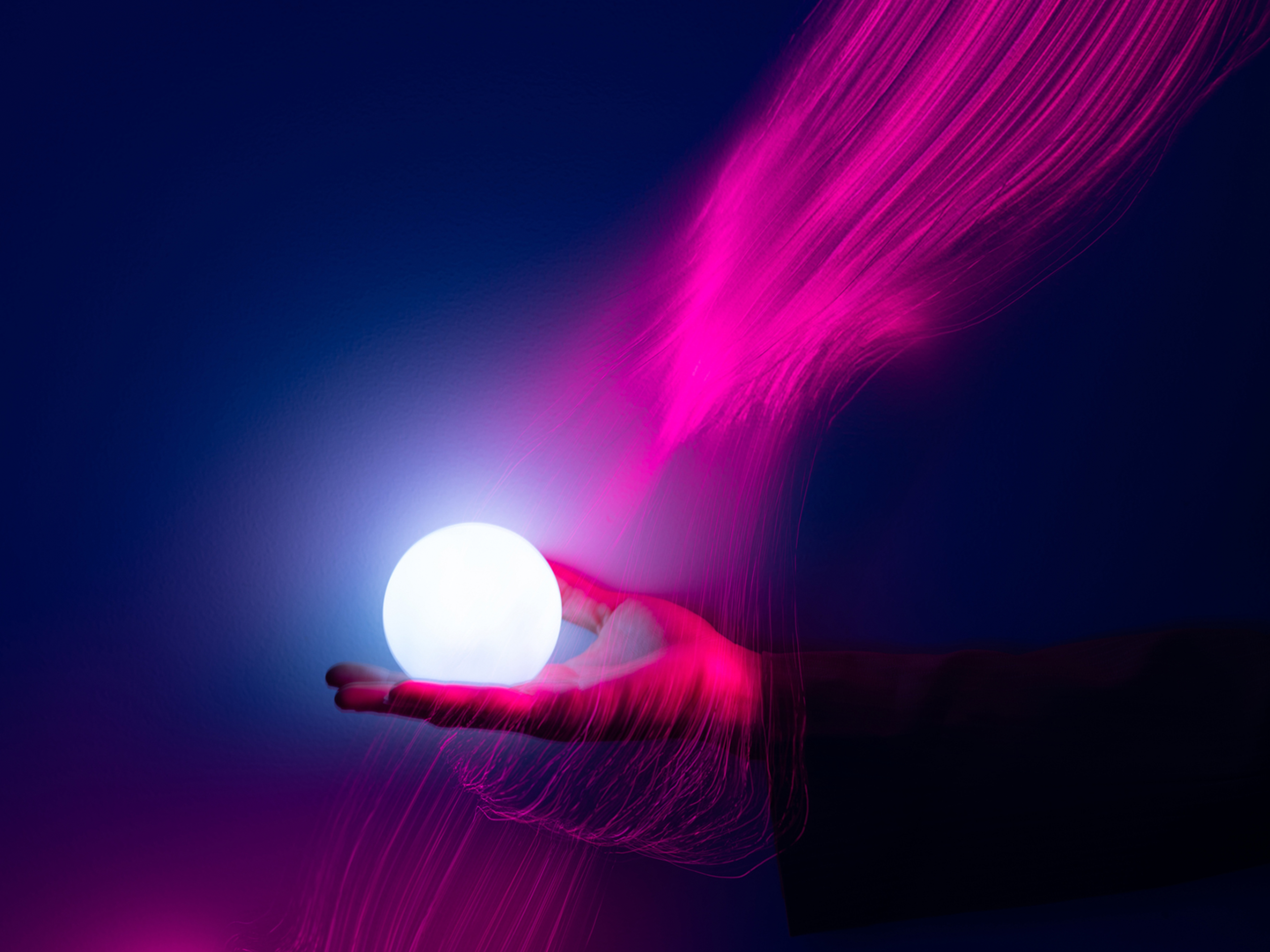 A hand holds a bright, glowing white orb, surrounded by vibrant magenta light trails and ethereal wisps against a deep blue and purple background.