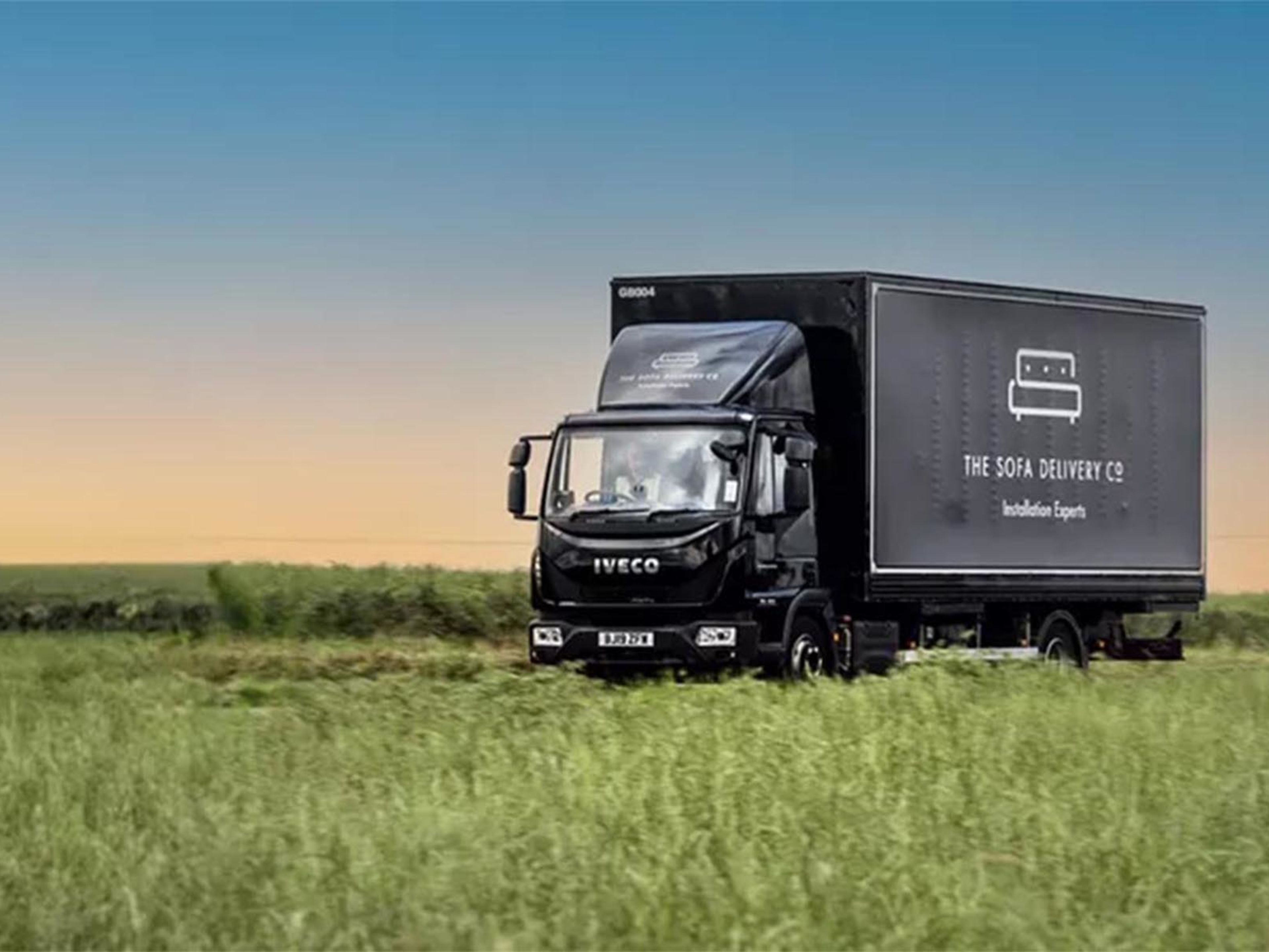 A black Iveco delivery truck with "The Sofa Delivery Co" logo on its side is parked in a wide green field under a blue and orange gradient sky.