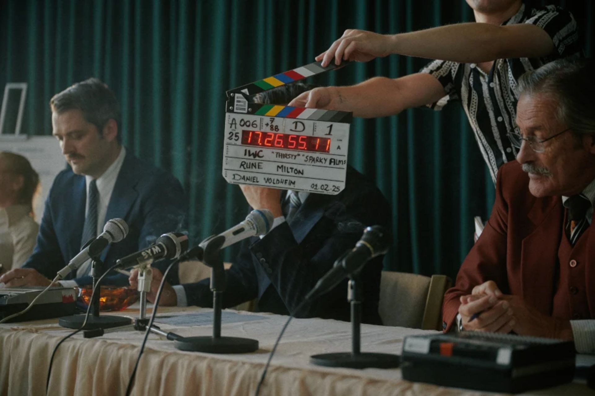 Behind-the-scenes film crew member holding a clapperboard with details for "Sparky Films" and director "Rune Milton" in front of actors seated at a retro press conference table with vintage microphones.