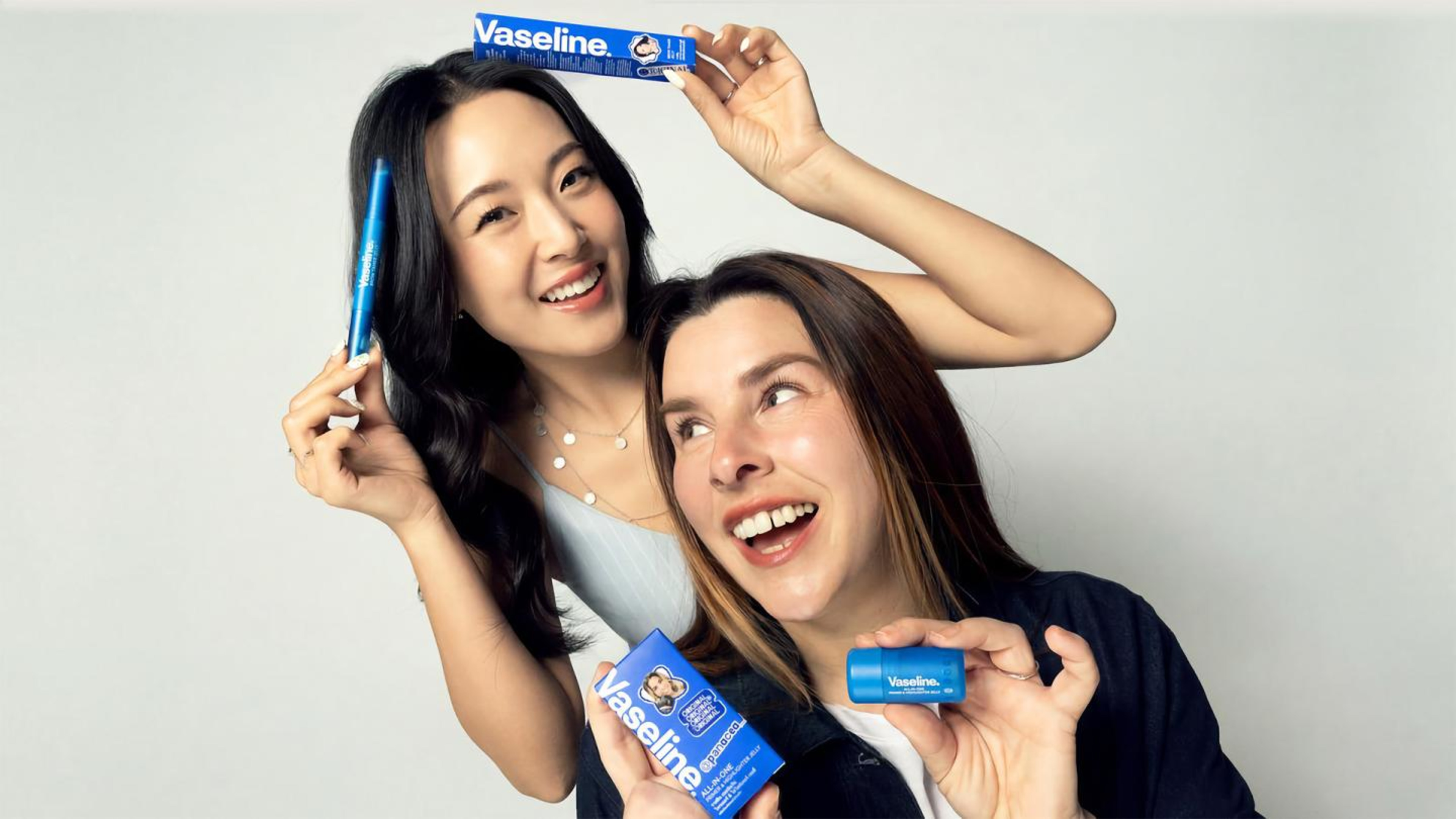 Two women smiling and posing playfully with Vaseline All-In-One skincare and highlighter jelly sticks.