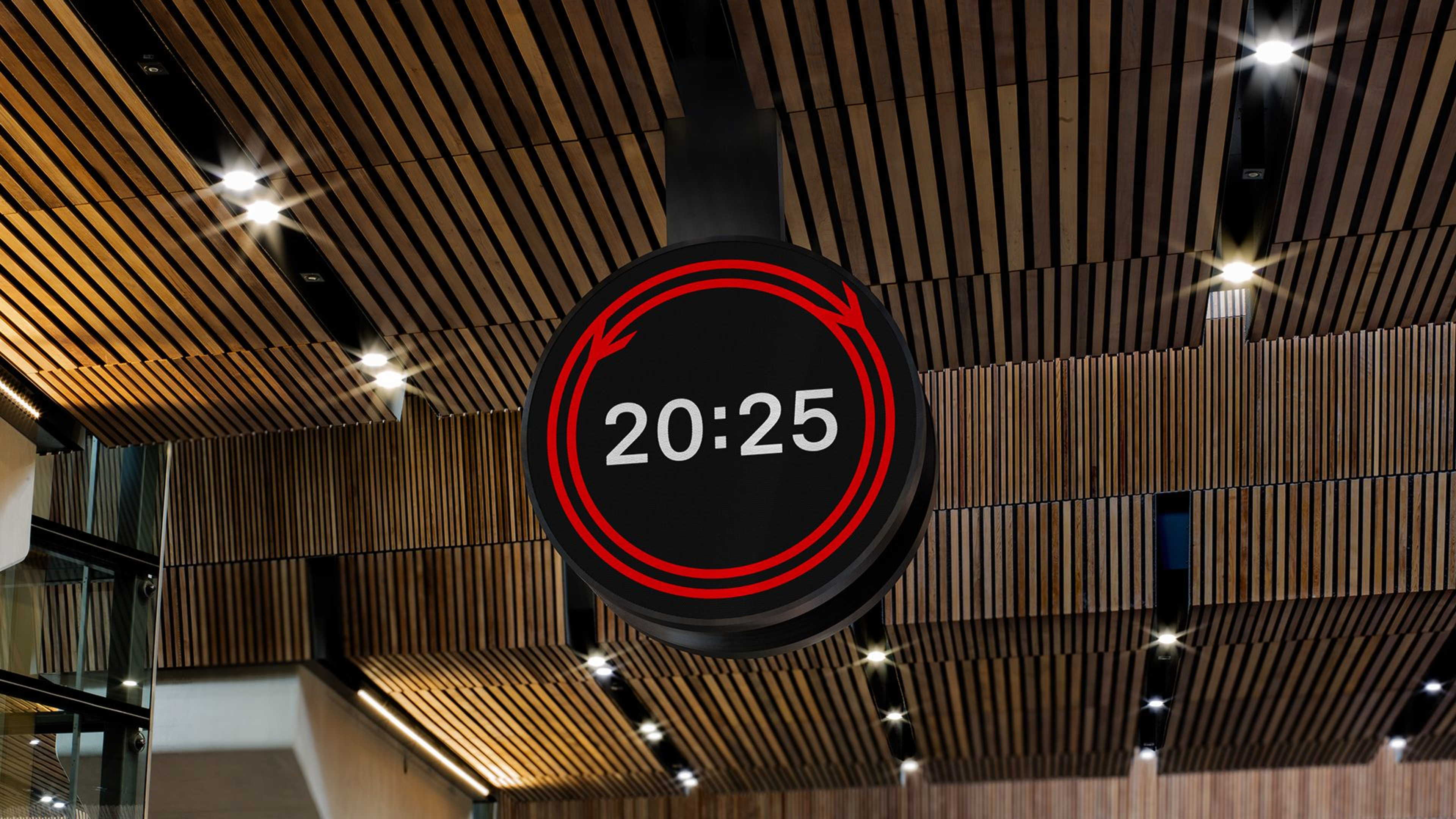 The circular Rail Clock hanging from a contemporary wooden-slatted station ceiling, featuring a digital display with a glowing red double-arrow graphic and white 24-hour time showing 20:25.