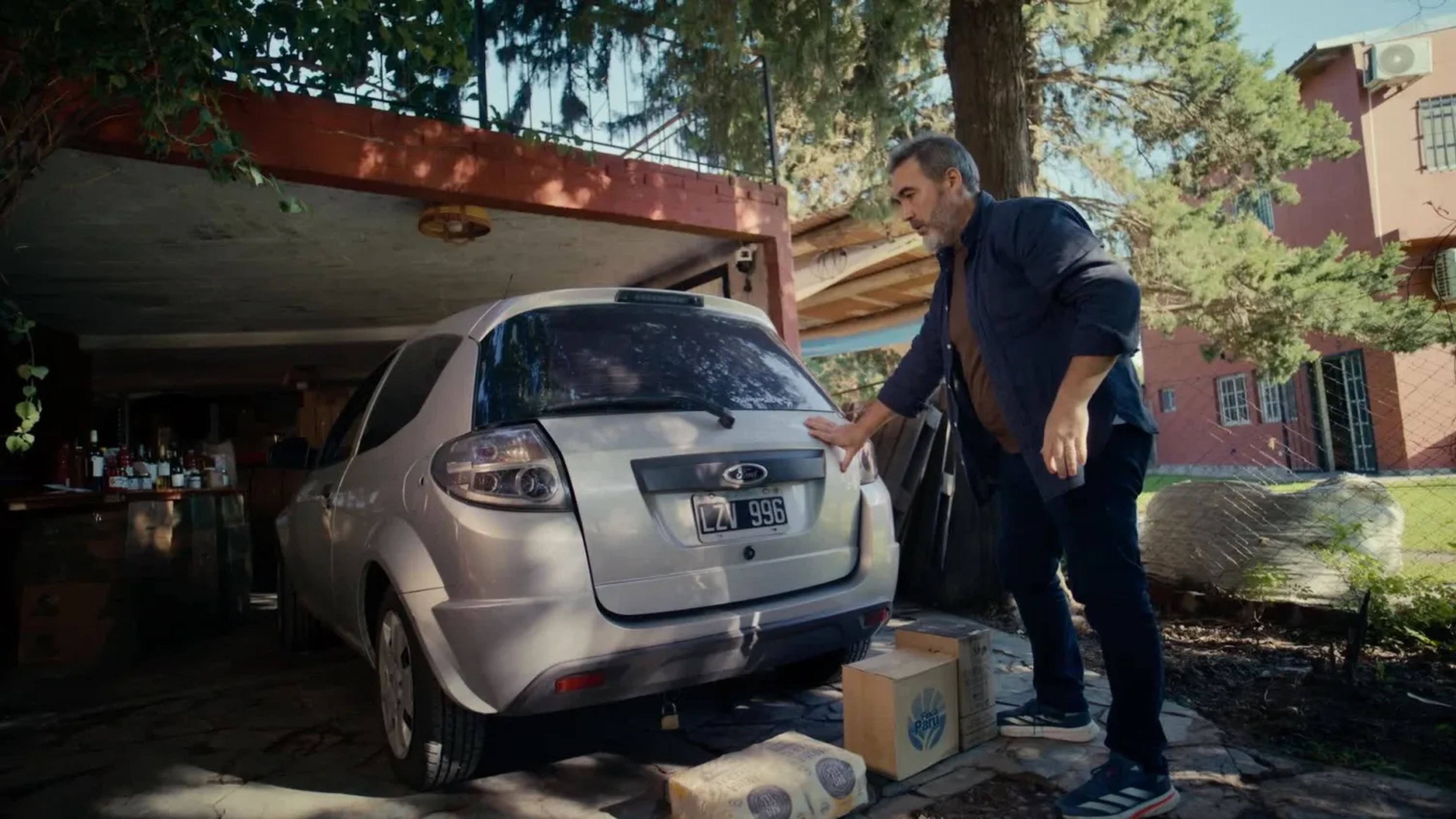 A small business owner in Argentina stands next to his silver hatchback car, looking at cardboard delivery boxes piled on his driveway and realising his vehicle is too small for peak season deliveries.