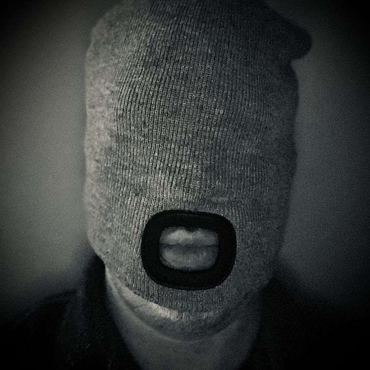 Close-up textured black and white photo of a person with a gray knit material covering their entire head and eyes, leaving only a mouth opening.