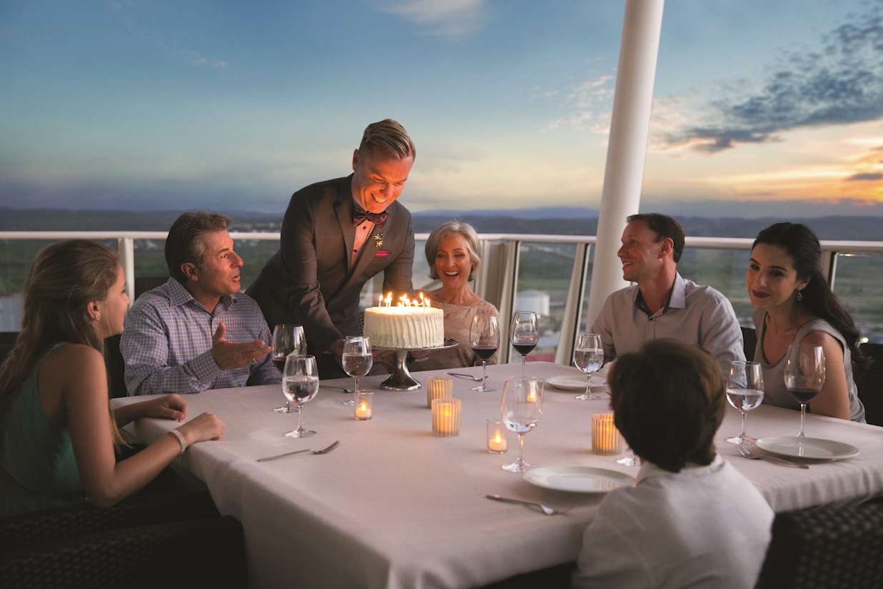 OA, Oasis of the Seas, Royal Suite Class, GENIE shoot, Royal Suite balcony dinner, Genie serving birthday cake for guests, service, family at dinner table, ocean and land view in background, early evening, twilight, sunset