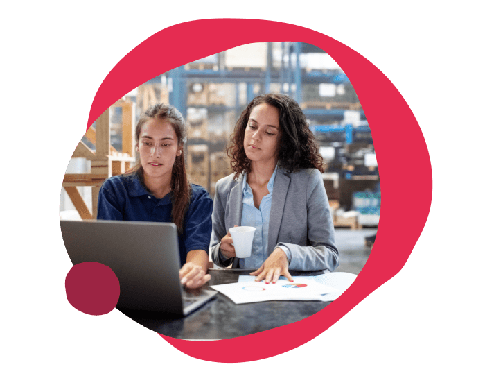working women discussing data analysis on laptop in warehouse, one holding a coffee cup, business meeting with charts and graphs