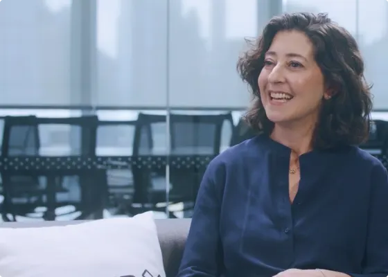 smiling woman in a blue shirt sitting on a couch with modern office background, conversational setting with natural lighting
