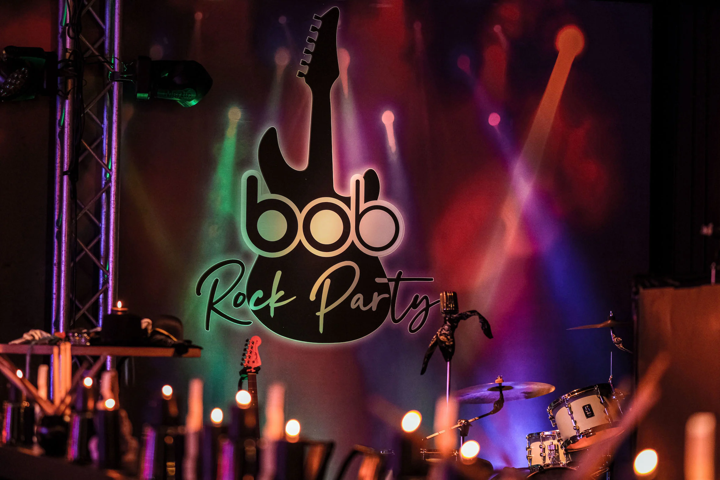 rock party backdrop with guitar silhouette, vibrant lights, candles, and drum set creating a lively atmosphere, colorful stage design featuring "bob Rock Party" logo and musical elements