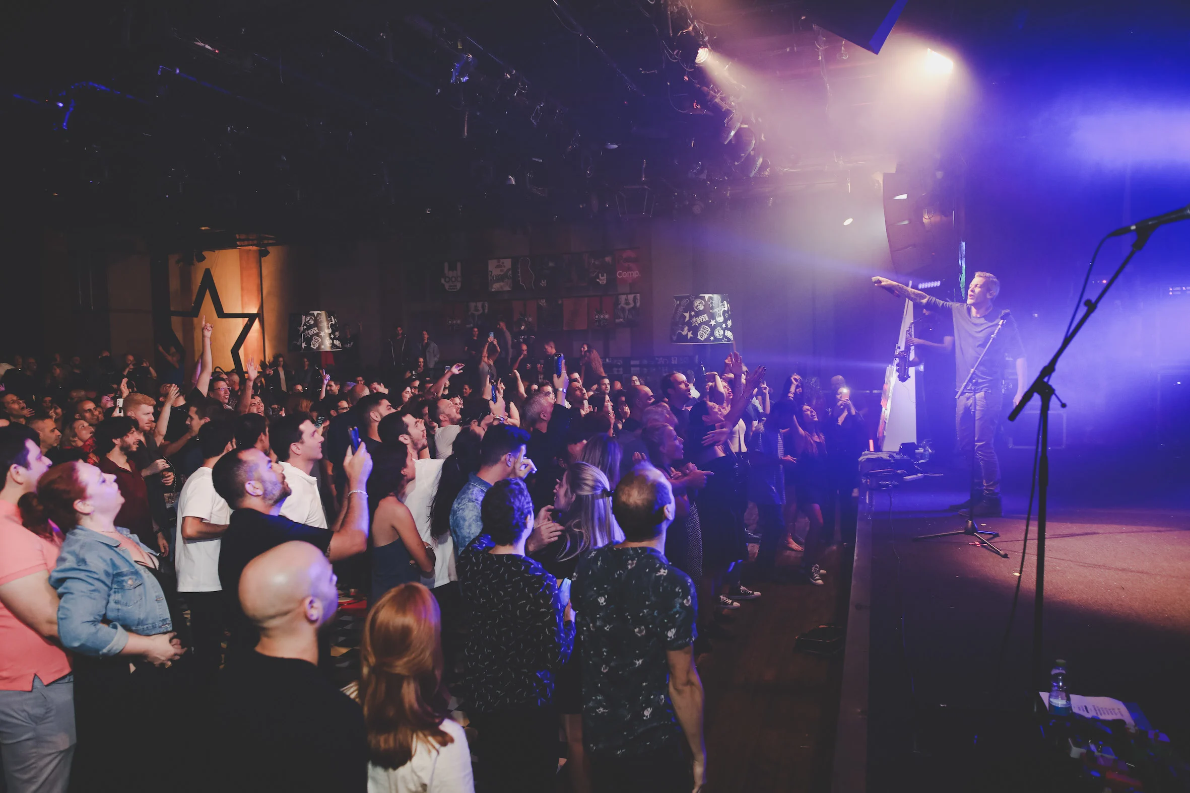 crowd at a live concert with raised hands and phones, performer engaging with audience under colorful stage lights, energetic live music event with fans enjoying the performance