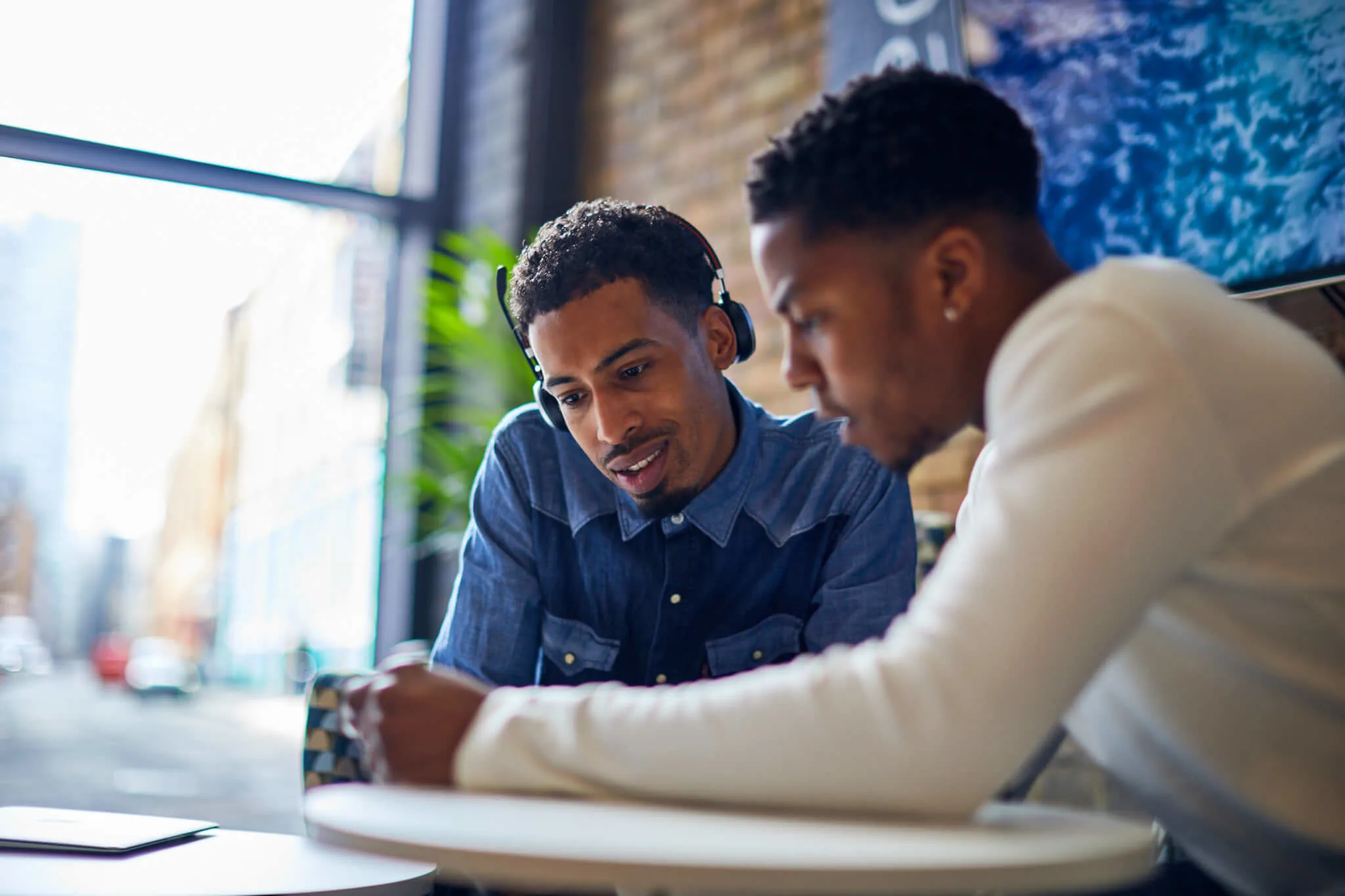 two young men collaborating over a device in a bright café, one wearing headphones, showing engagement and focus, café setting, teamwork, modern workspace