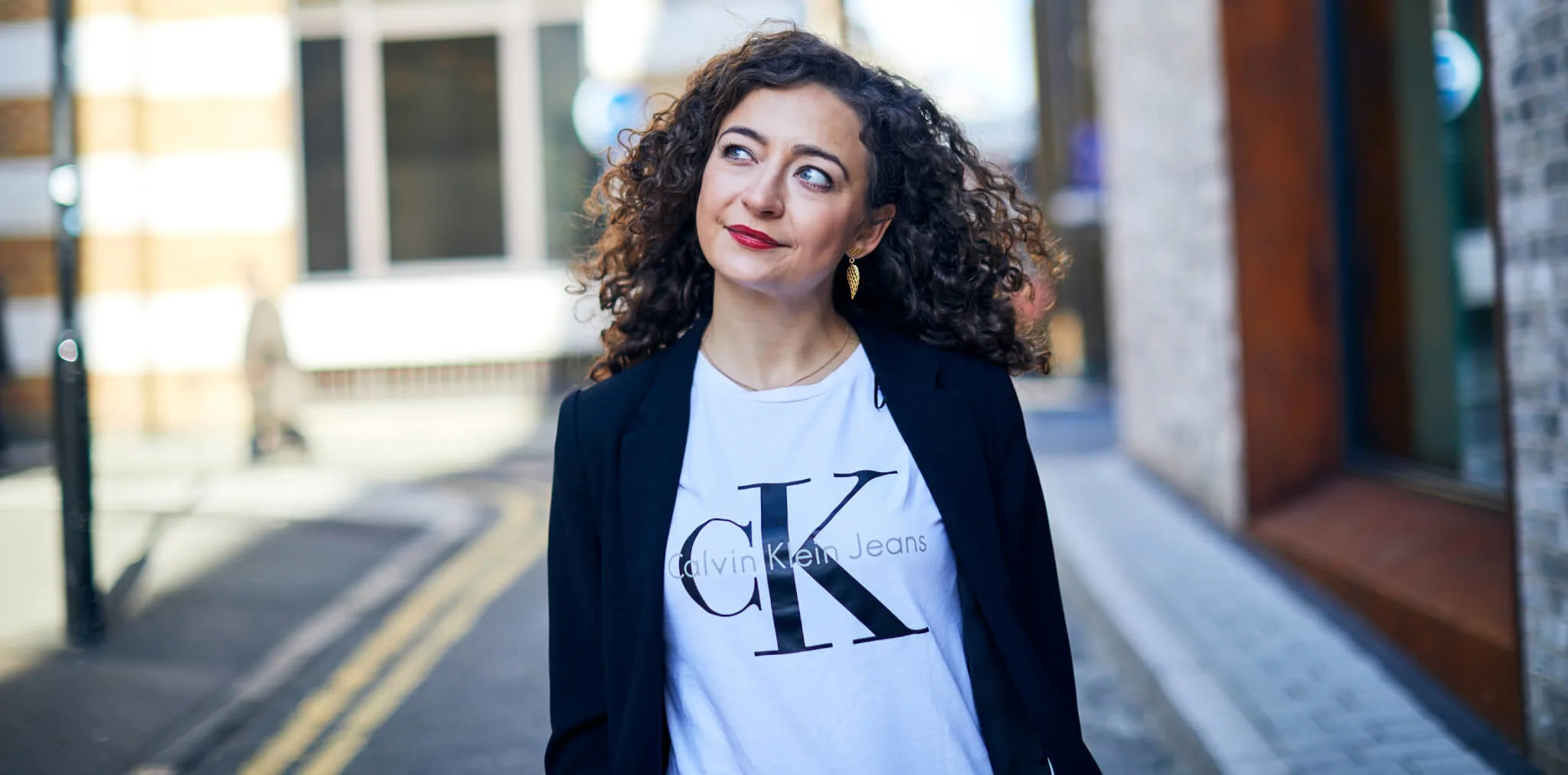 woman with curly hair in blazer and Calvin Klein t-shirt walking on street, bright urban background with blurred pedestrians