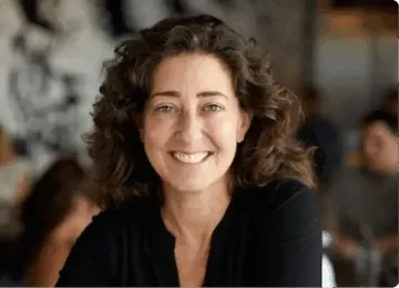 smiling woman with curly hair in a casual black shirt, sitting in a bright restaurant with blurred background, casual dining interior with patrons and artistic decor