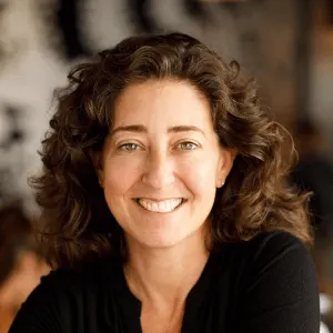 smiling woman with curly hair in a casual black shirt, natural light background with a warm, inviting atmosphere