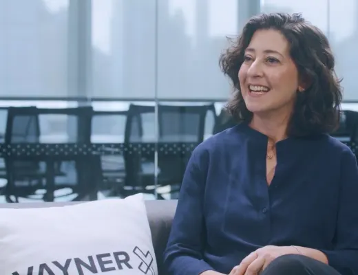smiling woman in blue shirt sitting on couch with WAYNER pillow, workplace backdrop, modern office setting, business interview atmosphere