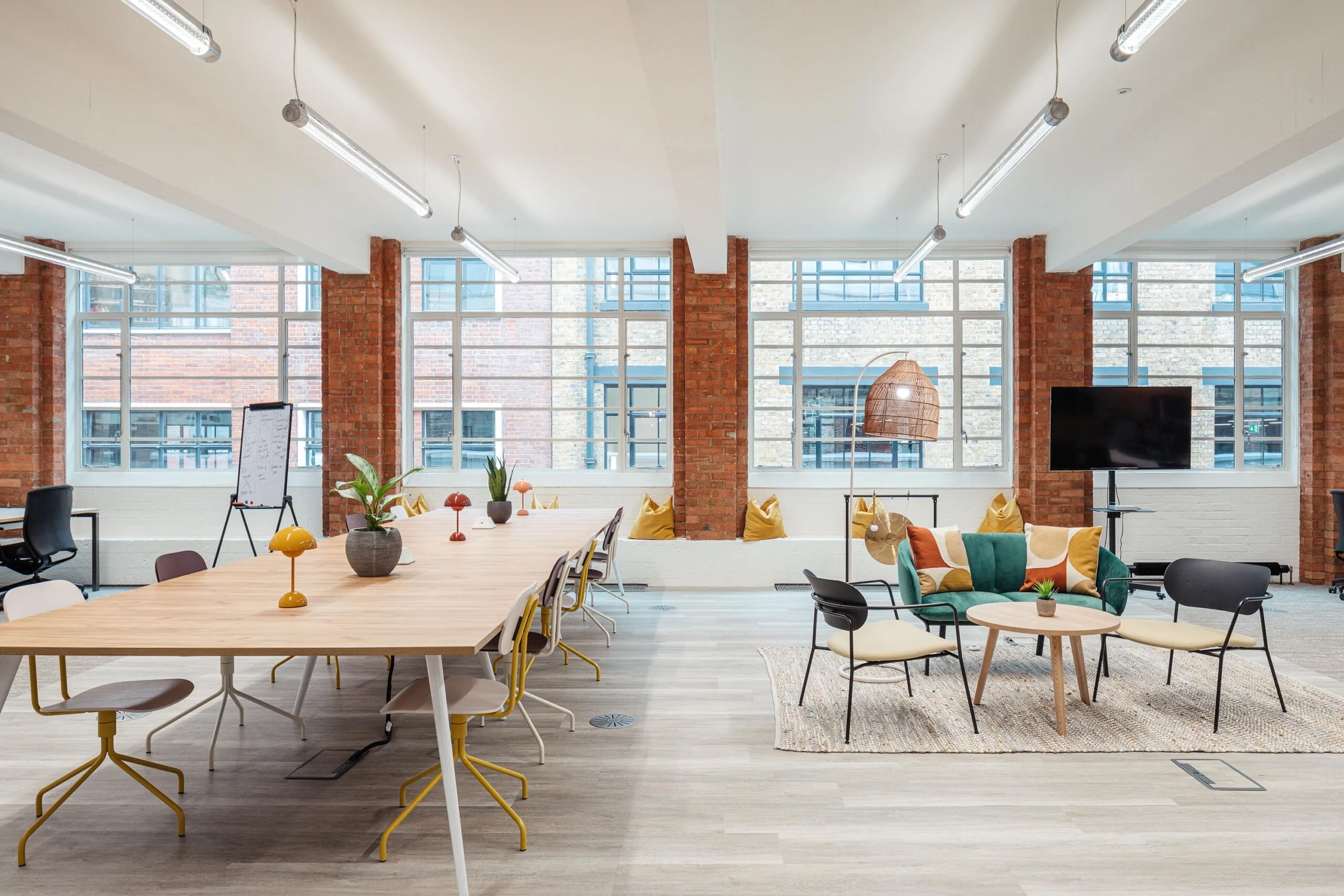 office interior with long wooden table, colorful seating area, natural light from large windows, and modern decor, stylish workspace with vibrant furnishings, plants, and a relaxed atmosphere