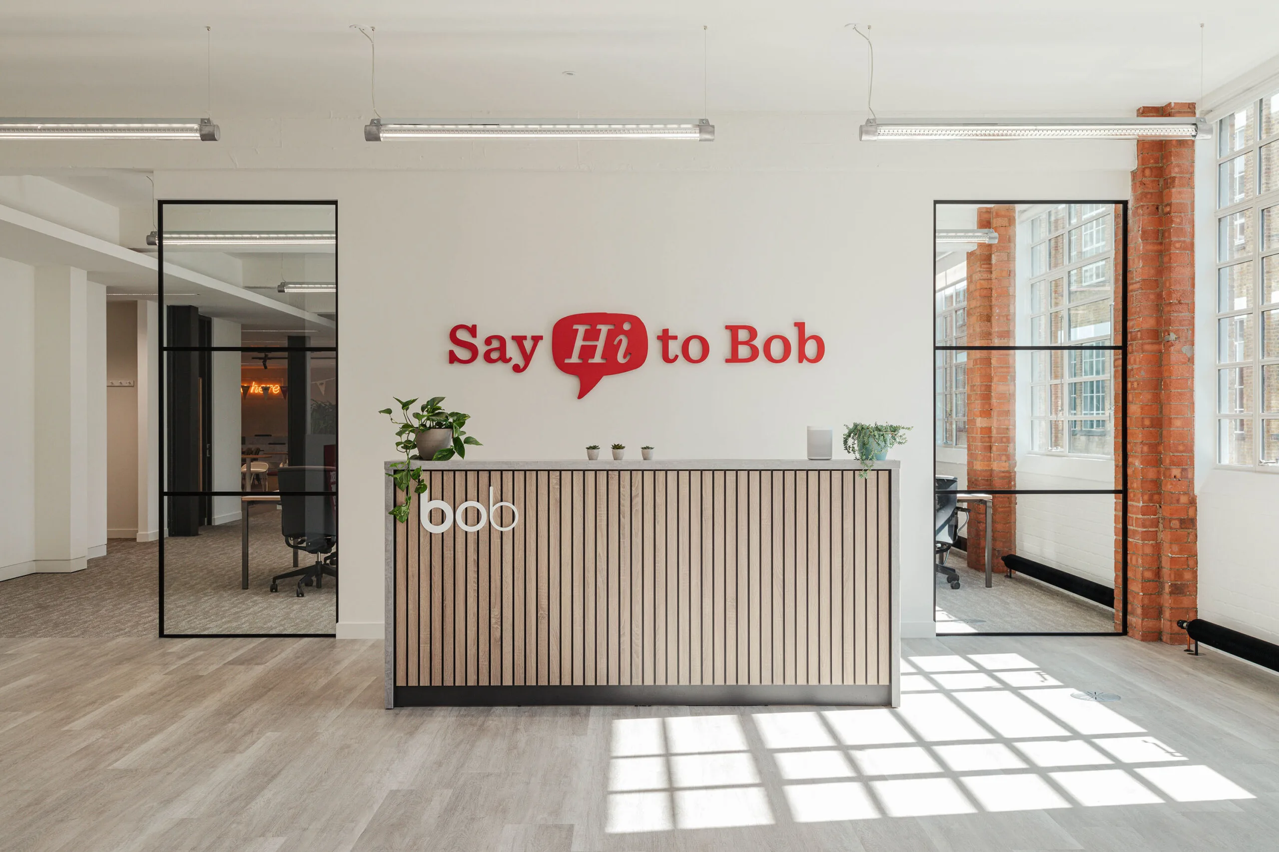 Say Hi to Bob, Modern office reception with wooden desk and potted plants, bright natural light through large windows.