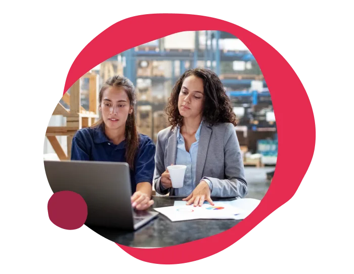 working women discussing data analysis on laptop in warehouse, one holding a coffee cup, business meeting with charts and graphs