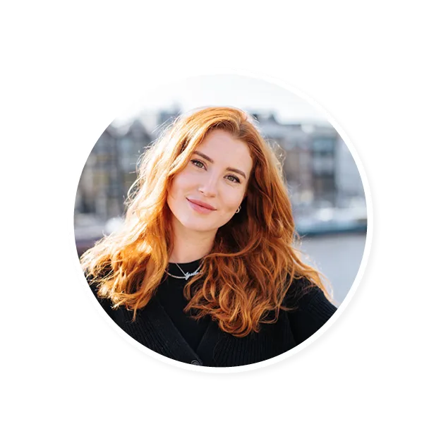 smiling woman with wavy red hair wearing a black sweater, sunlit cityscape background suggesting a cheerful atmosphere