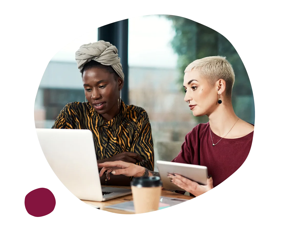 woman working on a laptop with a friend using a tablet in a modern workspace, two women collaborating in a bright office setting