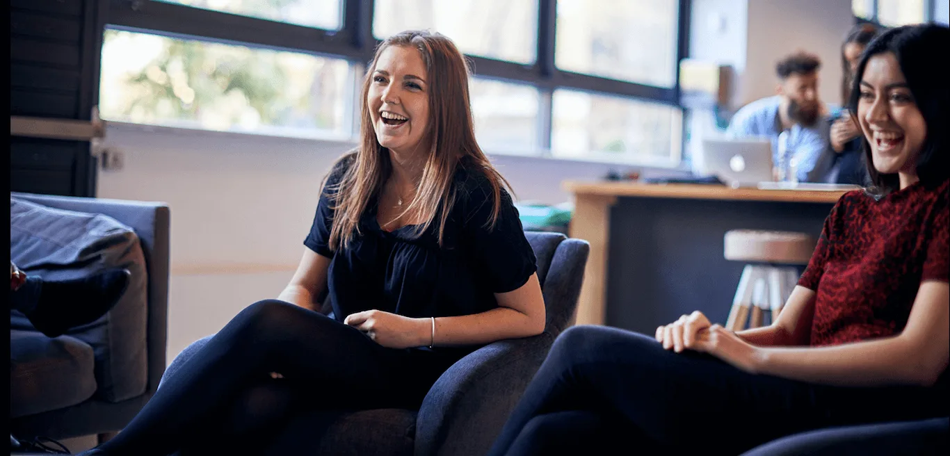 smiling women in a casual meeting, relaxed atmosphere, modern workspace, natural light, two friends enjoying conversation in an office setting