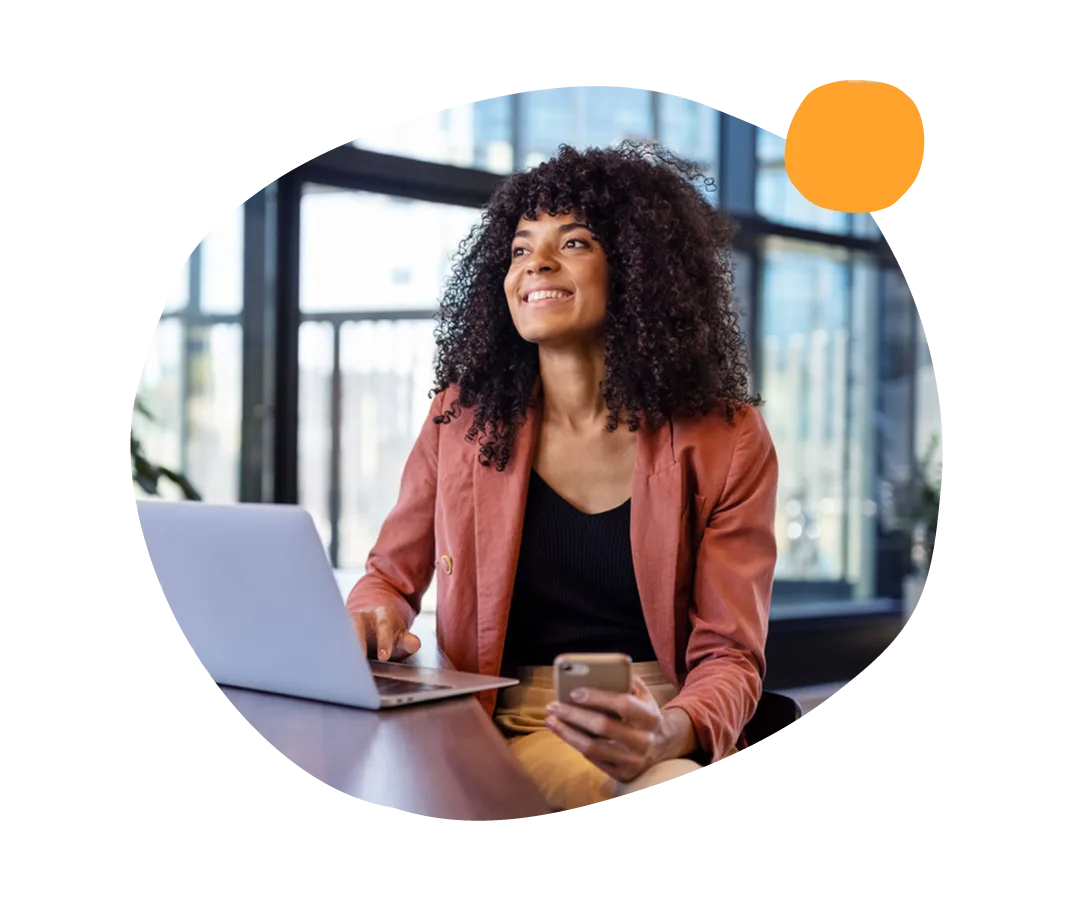 smiling woman using laptop and smartphone in modern office, curly hair and business attire in bright workspace