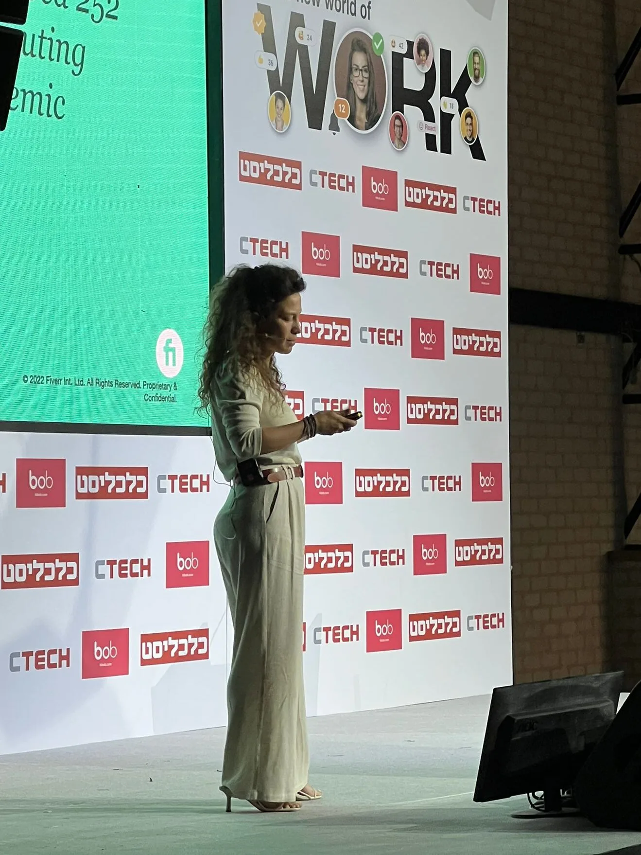 woman speaking on stage at a conference with a green presentation screen and various logos in the background, presentation on future work and technology trends