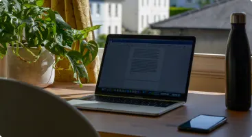 laptop-on-wooden-desk-with-notepad-and-phone, sunlit-window-view-with-green-plant-and-thermal-water-bottle