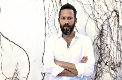 man in white shirt standing against a wall with vines, bearded man with crossed arms near greenery backdrop
