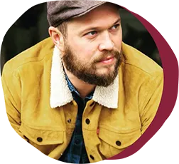 man with beard in mustard jacket and gray cap, close-up portrait in outdoor setting with warm color tones
